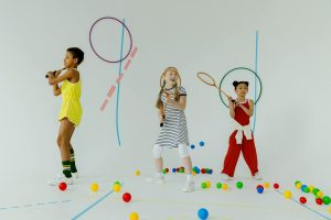 Three diverse children playing tennis indoors with rackets and colorful balls, showcasing fun and diversity.